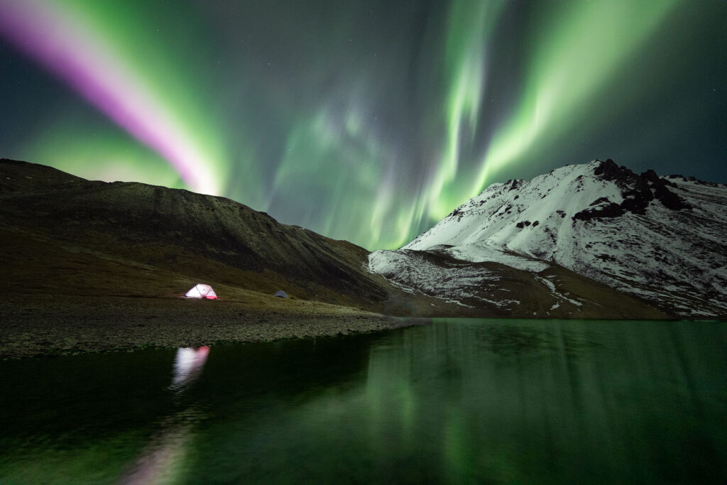 strong northern lights over lit tent and mountains