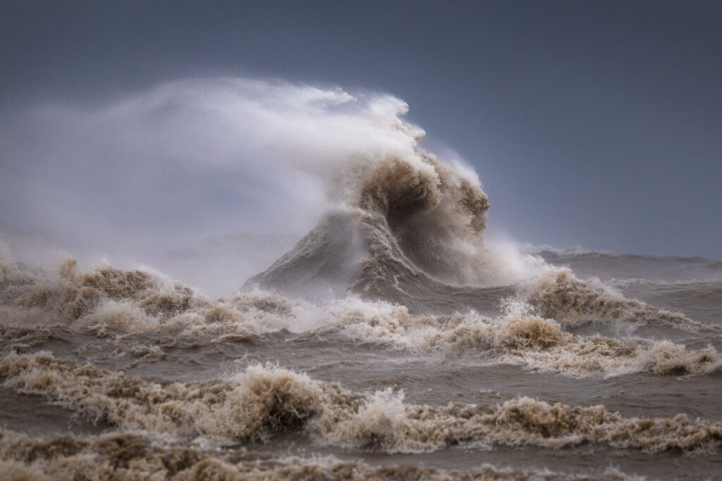 A large wave rolls forward, creating dramatic shapes in the wind