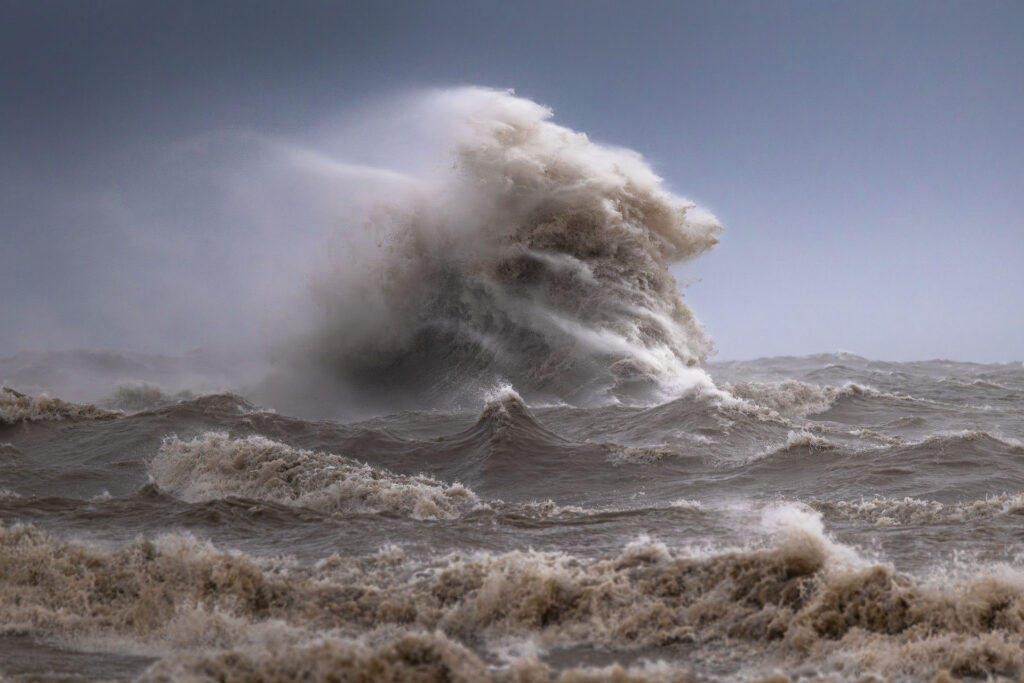 a powerful wave erupts and is blown apart by strong gusts of wind